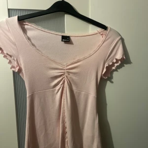 Ljusrosa topp från Gina Tricot - Supersöt ljusrosa topp med korta ärmar och v-ringad hals från Gina Tricot. Klänningen har snygg snörning framtill, volangkant på ärmarna och spetsdetaljer längs halsringningen. Mjuk och stretchig i bomullsmix, perfekt för dig som gillar romantisk stil.