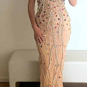 Champagnefärgad långklänning med stenar - Superglammig långklänning i champagnefärg med massor av färgglada stenar och glittriga detaljer. Klänningen är figurnära och har axelbandslös design. Perfekt för dig som vill sticka ut och glänsa på festen!