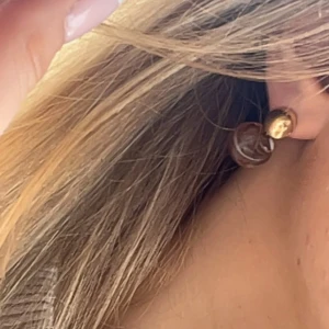 Guldiga örhängen med brun swirl - Snygga örhängen med rund, guldig plupp och hängande brun swirlad detalj i akryl. De har en cool, glansig finish och mixar guld med transparent brunt för en unik look. Perfekta för att ge din outfit lite extra edge.