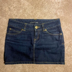 Mörkblå jeanskjol från Terranova - Säljer en mörkblå jeanskjol från H&M med klassisk femficksdesign och kontrastsömmar i gult. Kjolen har knapp och dragkedja framtill och är i kort modell. Perfekt för dig som gillar en enkel och cool denimstil.