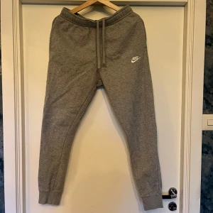 Grå mjukisbyxor från Nike - Gråa mjukisbyxor från Nike med broderad vit logga på benet. Byxorna har resår i midjan med snörning, muddar vid bensluten och en bakficka. Tillverkade i mjuk bomullsblandning för maximal komfort.