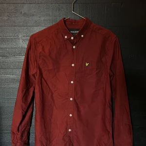 Vinröd skjorta (Lyle & Scott) - Snygg vinröd skjorta från Lyle & Scott med klassisk button-down krage och vita knappar. Skjortan har långa ärmar och en liten gul broderad logga på bröstet.