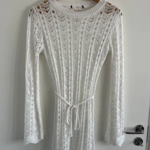 Vit stickad långklänning med knyt - Superfin vit långklänning i stickat material med vackert spetsmönster. Klänningen har långa ärmar, rund halsringning och ett knytband i midjan. Baksidan har en snygg öppen rygg för extra detalj. Perfekt för sommarens alla tillfällen!