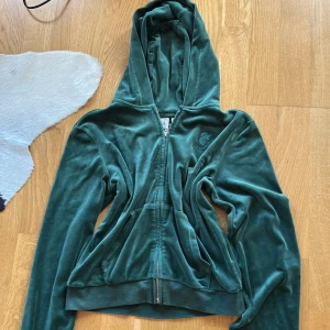 Grön velour hoodie Juicy Couture - Säljer en grön hoodie från Juicy Couture i mjuk velour med broderad JC-logga på bröstet. Tröjan har huva, dragkedja framtill och två fickor. Jätte mjuk .Passformen är normal och ärmarna är långa.