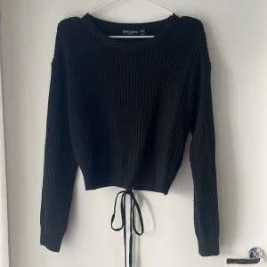 Svart stickad tröja med snörning - Svart stickad tröja från Brave Soul med långärmad design och  snörning i ländrygg Tröjan har ett öppet  ryggslut  med snygg knyt-detalj som ger en unik look. Perfekt för dig som gillar trendiga och bekväma plagg med lite edge.