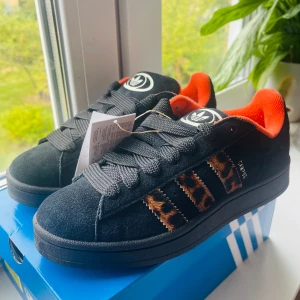Adidas Campus svart/leopard sneakers - Svarta Adidas Campus sneakers i mocka med ikoniska tre ränder i leopardmönster och cool orange insida. Tjocka svarta snören och klassisk Campus-logga på sidan. Perfekta för dig som vill sticka ut med en djärv och trendig look.