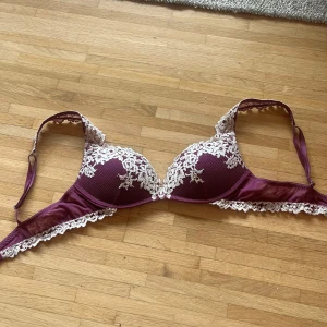 Vinröd spetsdetaljerad korsett-topp - Säljer en vinröd korsett-topp med vackra vita spetsbroderier och blommönster över kuporna. Toppen har justerbara axelband och fina spetskanter längs nederkanten. Materialet är mjukt och glansigt med en lyxig känsla, perfekt för att lyfta din outfit.