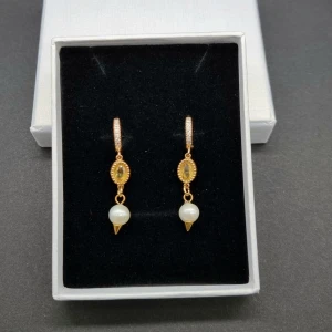Guldiga örhängen med pärlor och stenar - Snygga örhängen i guldton med små gnistrande stenar på ringen, oval gul sten i mitten och en vit pärla som hänger längst ner. Perfekt för dig som gillar eleganta detaljer och vill sticka ut lite extra.