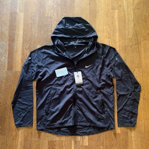 Svart Nike vindjacka  - Nike vindjacka i svart med huva och hel dragkedja framtill. Jackan har en stilren look med Nike-logga på bröstet och 'Nike Running' detaljer på insidan. Lätt och smidig polyester, perfekt för aktiva dagar. Loose fit för skön rörelsefrihet.