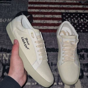 Saint Laurent vita sneakers i canvas - Snygga vita sneakers från Saint Laurent med handskriven logga på sidan och präglad text på hälen. Skorna har rund tå, platt sula och snörning. Tillverkade i canvas med detaljer i skinn, perfekt för dig som gillar minimalistisk och clean stil. Storlek 42 men passar 41 ochså