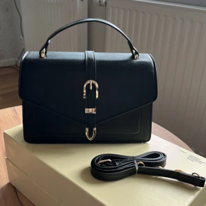 Svart handväska med gulddetaljer - Stilren svart handväska med elegant guldfärgad spänne framtill. Väskan har en klassisk rektangulär form, topphandtag och avtagbar axelrem. Materialet är slätt och ger en lyxig känsla. Perfekt för dig som gillar minimalistisk design och snygga detaljer. HELT NY🤩