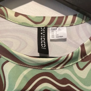 Grönbrun mönstrad klänning H&M Divided - Cool kortklänning från H&M Divided med grönt och brunt vågigt mönster. Klänningen har korta ärmar, rund hals och snygga knyt-detaljer i midjan. Bra skick!