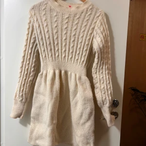 Stickad vit kortklänning med kabelmönster - Supersöt stickad klänning i vit ton med klassiskt kabelstickat mönster och rund hals. Modellen är kort med markerad midja och långa ärmar. Den har någon fläck jag inte riktigt vet va de är men jag antar att de nagellack 