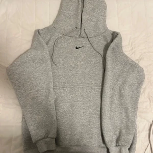 Grå Nike hoodie med huva, storlek M - Klassisk grå hoodie från Nike med svart broderad swoosh på bröstet. Tröjan har huva med snörning, ribbade muddar och en skön, mjuk insida. Perfekt för chill dagar eller när du vill ha en enkel streetstyle-look.