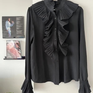 Svart volangskjorta med plisserad krage - Svart skjorta med plisserad volang längs knäppningen och en bred plisserad krage. Långa ärmar med plisserade manschetter ger en cool och unik look. Skjortan har klassisk knäppning framtill.