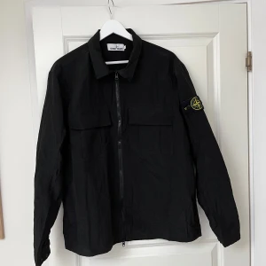 Svart overshirt från Stone Island - Svart overshirt från Stone Island med klassisk krage, två stora bröstfickor och dragkedja framtill. Ikonisk logga på vänster ärm. Lätt och stilren jacka i syntetmaterial, perfekt för lager-på-lager och streetwear-vibe. Använd 4-5 gånger. Nypris 2999. 