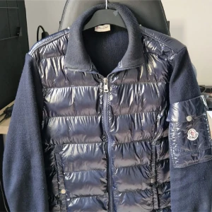 Mörkblå pufferjacka från Moncler - Snygg mörkblå pufferjacka från Moncler med glansig quiltad front och ärmar i stickad ull. Jackan har dragkedja framtill, hög krage och en ikonisk Moncler-logga på ärmen. Perfekt för dig som vill ha en stilren och exklusiv look.