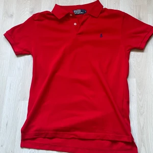 Röd pikétröja från Ralph Lauren - Snygg röd pikétröja från Polo Ralph Lauren med klassisk krage och två knappar framtill. Tröjan har korta ärmar och den ikoniska broderade loggan på bröstet. Perfekt för en clean och sportig look.