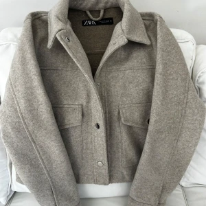 Beige jacka från Zara - Felfri, knappt använd beige jacka från Zara i en stilren design. Jackan har en klassisk krage och knappar framtill. Två stora fickor med lock på framsidan ger en cool look. Perfekt för våren som kommer nu!