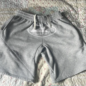 Grå sweatshorts med snörning - Snygga grå sweatshorts med elastisk midja och snörning framtill. Shortsen har en vit tryckt detalj på framsidan och praktiska fickor på sidorna samt en bakficka med lock. Perfekta för chill dagar eller träning.