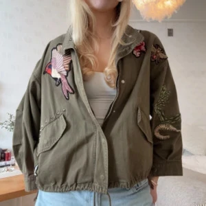 VIRALA slutsålda H&M jackan - Snygg olivgrön oversized parkajacka med broderade detaljer som rosor, orm och fågel. Jackan har dragkedja, stora fickor, snörning i midjan och nitar på ena fickan. Perfekt för dig som vill sticka ut med coola detaljer och relaxed fit.