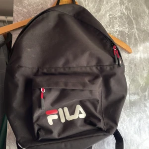 Svart ryggsäck från FILA - Klassisk svart ryggsäck från FILA med stort huvudfack och en praktisk ytterficka framtill. Väskan har röda detaljer på dragkedjorna och tydlig FILA-logga i vitt och rött på framsidan. Tillverkad i slitstark polyester, perfekt för skola eller vardag.