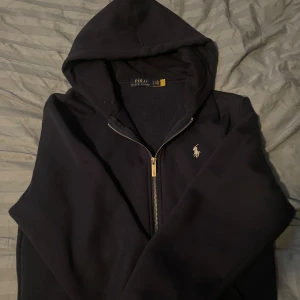 Mörk blå hoodie från Polo Ralph Lauren - Mörk blå hoodie från Polo Ralph Lauren med klassisk logga broderad på bröstet. Tröjan har huva, dragkedja i metall och är långärmad. Tillverkad i mjuk bomull som känns skön mot huden. Perfekt för chill dagar och streetwear-stil. Den är i storlek M men skulle säga att den passar folk som har S mer!
