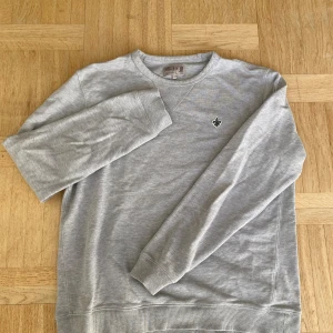 Grå sweatshirt från Morris - Snygg grå sweatshirt från Morris i storlek S. Tröjan har ett litet broderat emblem på bröstet och klassisk rund hals. Tillverkad i mjuk bomull med ribbade muddar vid ärmslut och nederkant. Perfekt för chill dagar eller när du vill vara casual.