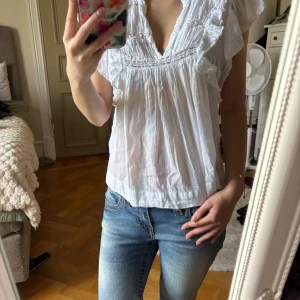 Vit blus med volang från Neo Noir - Superfin vit blus från Neo Noir med v-ringning, volangdetaljer längs axlar och halsringning samt lätt rynkade partier. Luftig och flowy passform, perfekt för varma dagar. Tillverkad i tunn bomull som känns skön mot huden. Skriv för fler bilder eller om du har en fråga🩷