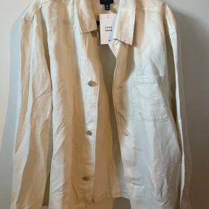 Beige linne overshirt från H&M - Snygg beige skjorta från H&M i linnemix, perfekt för dig som gillar en clean och chill stil. Skjortan har klassisk krage, lång ärm, knappar framtill och en bröstficka. Passar dig som vill ha en luftig och enkel look.