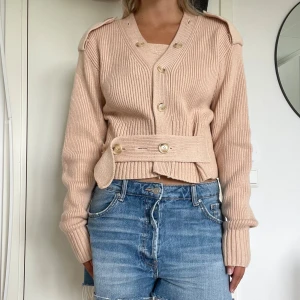 Beige ribbad kofta med bälte - Säljer en beige ribbad kofta med v-ringning och snygga knappar framtill. Koftan har ett brett bälte med knappar i midjan och markerade axlar med dekorativa knappar. Långärmad och croppad modell som ger en cool och trendig look.