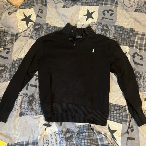 Svart stickad tröja Polo Ralph Lauren - Svart stickad tröja från Polo Ralph Lauren med ribbad krage och knappdetaljer vid halsen. Klassisk vit logga broderad på bröstet. Långärmad och med ribbade muddar vid ärmslut och nederkant. Perfekt för dig som gillar stilren och enkel design. Lite nopprig men annars är den i väldigt bra skick.