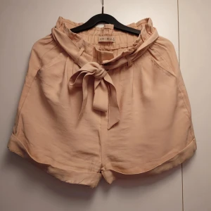 Beige shorts med knyt från Amisu - Snygga beige shorts från Amisu med hög midja och bred knyt i midjan. Modellen har papperspåse-midja och uppvikta benslut för en chill vibe. Materialet känns mjukt och lätt, perfekt för varma dagar. Passar dig som gillar en loose fit och vill ha något stilrent men ändå trendigt.