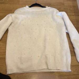 Vit stickad tröja med pärlor - Superfin vit stickad tröja med pärldetaljer framtill. Tröjan har rund halsringning, långa ärmar och ribbade muddar vid ärmslut och nederkant. Perfekt för dig som gillar en clean look med lite extra bling.