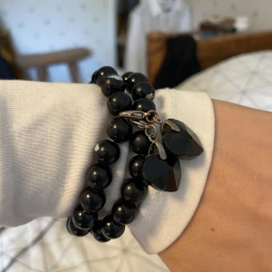 Svart pärlarmband med hjärtberlocker - Snyggt svart armband med glansiga pärlor och två svarta hjärtformade berlocker. Armbandet är elastiskt och har två band som sitter ihop. Perfekt accessoar för att lyfta din outfit med en touch av svart.