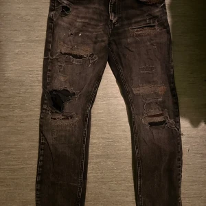 Svarta ripped jeans från Zara - Säljer ett par svarta jeans från Zara med snygga slitningar och hål på benen för en edgy look. Jeansen har smal passform och klassisk femficksdesign. Perfekta för dig som gillar streetstyle och vill sticka ut lite extra.