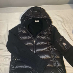 Svart Moncler cardigan - Säljer en svart dunväst från Moncler med stickade ärmar och detaljer. Västen har en quiltad, glansig framsida, huva och dragkedja. På ena ärmen finns en ficka med Moncler-logga. Perfekt för lager-på-lager och riktigt snygg streetstil.