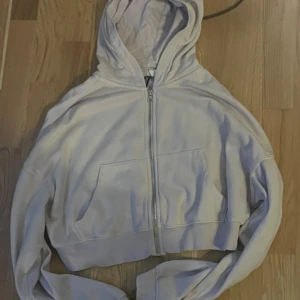 Ljusgrå croppad hoodie med dragkedja - Säljer en ljusgrå croppad hoodie med dragkedja framtill och stor huva. Tröjan har långa ärmar och en rymlig magficka. Perfekt för en chill och avslappnad stil.