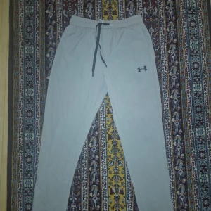 Under Armour ljusgrå träningsbyxor - Snygga ljusgrå träningsbyxor från Under Armour med elastisk midja och svart snörning. Byxorna har en slim passform och är gjorda i ett lätt syntetmaterial med HeatGear-teknologi som håller dig sval. Perfekta för gymmet eller sportaktiviteter.