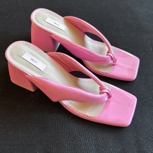 Rosa klackade sandaler från Nelly - Snygga rosa sandaler från Nelly med fyrkantig tå och bred klack. Skorna har en glansig finish och trendig design med knutdetalj framtill.