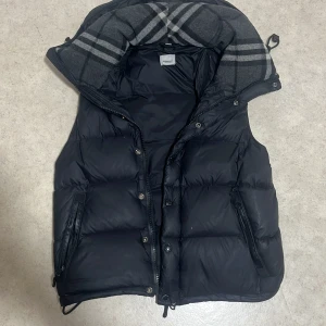 Svart dunväst från Burberry - Snygg svart dunväst från Burberry med klassiskt rutigt foder i huvan. Västen har dragkedja och knappar framtill, två fickor med dragkedja och justerbar nederkant. Perfekt för lager-på-lager och riktigt stilren look. Västen har även med armarna som kan sättas på passar perfekt för vintern när de är kyligt😊