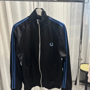 Svart Fred Perry hoodie med blå ränder - Svart hoodie från Fred Perry med blå ränder längs ärmarna och vit dragkedja. Klassisk hög krage och broderad logga på bröstet. 