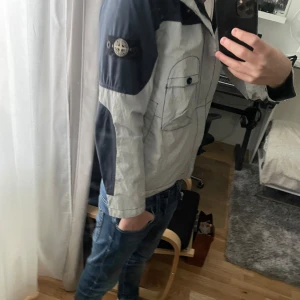 Stone Island vindjacka med huva - Säljer en fet vindjacka från Stone Island i ljusgrått med mörkblå detaljer på ärmar och axlar. Den har riktigt cool reflex som syns när man tar bild med flash på kameran (se bild 5) Jackan har huva, dragkedja och ikonisk patch på vänster ärm. Junior stl men passar som xs. Bra skick och självklart äkta(kvitto finns) riktigt fet och stilren jacka till hösten.