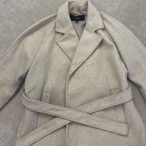Beige kappa från Vero Moda med bälte - Snygg beige kappa från Vero Moda med klassisk krage och bälte i midjan. Jackan har en stilren look, är helfodrad och har raka linjer. Perfekt för dig som gillar en clean och tidlös vibe. Knappt använd 