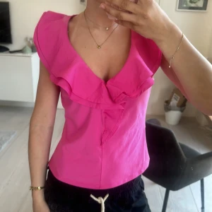 Rosa volangtopp - Säljer en snygg rosa topp med volangdetaljer och djup v-ringning. Toppen har en feminin och trendig look, perfekt för att sticka ut. Materialet känns lätt och bekvämt. Helt oanvänd