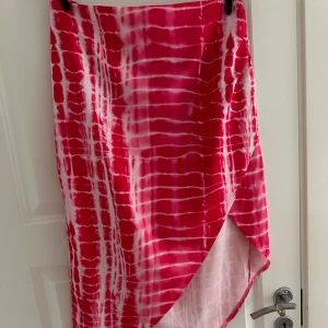 Rosa och vit omlottkjol i satinlook - Snygg långkjol i rosa och vitt med batikmönster och omlott-design. Kjolen har en lätt glansig satinliknande yta och en cool asymmetrisk slits framtill. Perfekt för dig som vill sticka ut med färg och mönster.