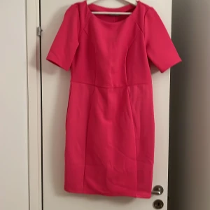 Kort rosa klänning med kort ärm - Säljer en snygg kort klänning i stark rosa färg. Klänningen har rund halsringning och korta ärmar, med en figurnära passform som ger en stilren siluett. Perfekt för dig som gillar färg och vill sticka ut lite extra.