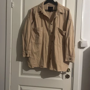 Beige overshirt från Lager 157 - Snygg beige overshirt från Zara med klassisk krage, stora fickor framtill och knappar hela vägen. Materialet är mjuk bomull och modellen är loose fit med slits på sidorna. Perfekt att slänga över en t-shirt för en chill look.