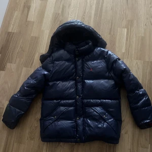 Mörkblå dunjacka från Polo Ralph Lauren - Säljer en mörkblå dunjacka från Polo Ralph Lauren med röd logga på bröstet. Jackan har huva, guldfärgade knappar och två stora fickor framtill. Barnstorlek xl men passar en S. 