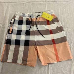 Burberry rutiga shorts med snörning - Snygga shorts från Burberry med klassiskt rutigt mönster i beige, svart, vit och röd. De har elastisk midja med svart snörning och är tillverkade i ett lätt syntetmaterial. Perfekta för sommaren och har fickor på sidorna för extra funktion.